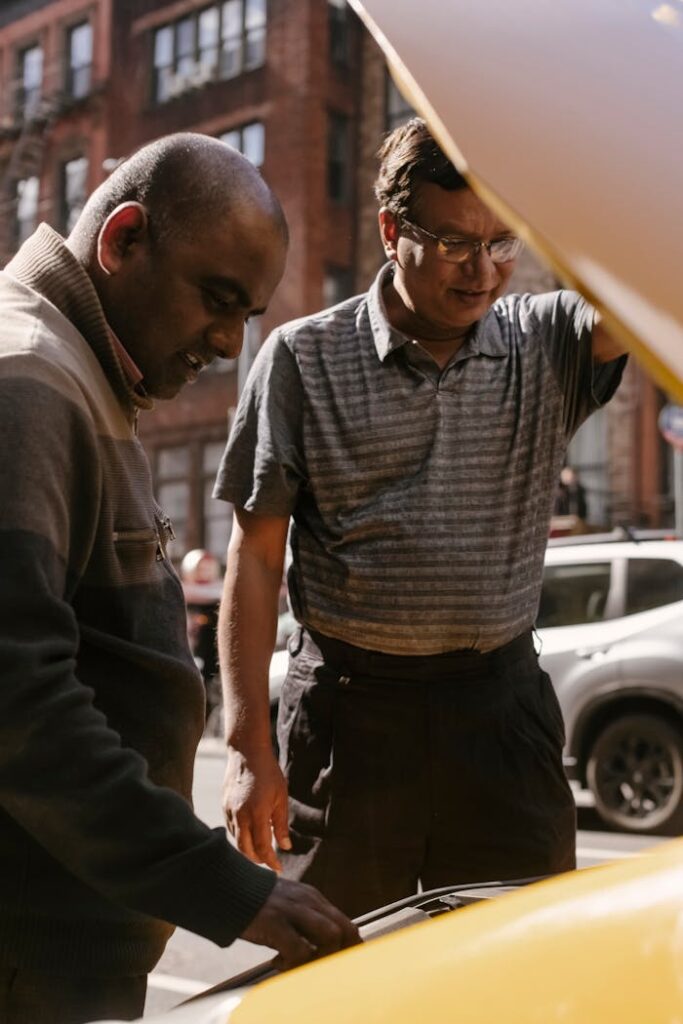 Two men inspect a car engine on a city street, seeking a solution to car trouble.